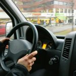 Hands on a steering wheel driving a van through an urban area with shops visible outside.