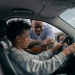 Young boy sitting at the wheel while father instructs from outside, learning to drive.